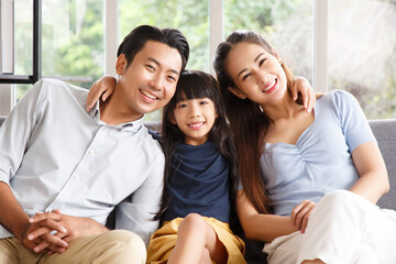 Obraz premium Portrait of a happy young family. Mom, dad and daughter look at the camera and smile. The faces of Asian parents and their child in living room.