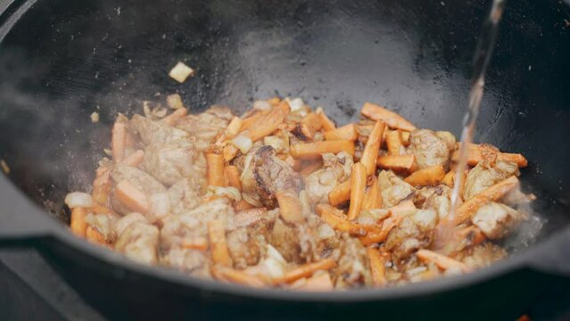 Water is poured into the cauldron where onions and meat and carrots are fried. Close-up.