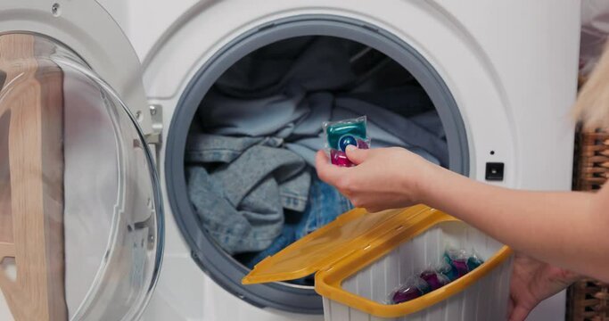 The woman sits at the washing machine and performs household chores. The clothes placed in the drum are waiting for the program to start. The girl holds a box of capsules drops one inside the laundry.