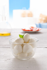 morsels of mozzarella in a glass bowl, with slices of tomato and olive oil placed on a white wooden table