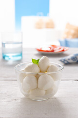 morsels of mozzarella in a glass bowl, with slices of tomato and a glass of water placed on a white wooden table
