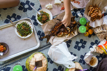 the tradition of eating together, seafood, grilled fish, fried prawns, vegetable stir-fried kangkung, young coconut, with their hands taking the grilled fish side dish, on a plastic mat.