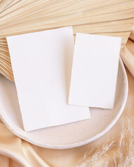 Blank cards on plate on dried palm leaf and beige silky fabric close up, greeting or wedding mockup