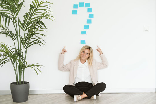 Businesswoman sitting on the floor in office pointing at question mark above her