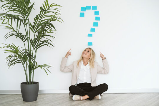 Businesswoman sitting on the floor in office pointing at question mark above her