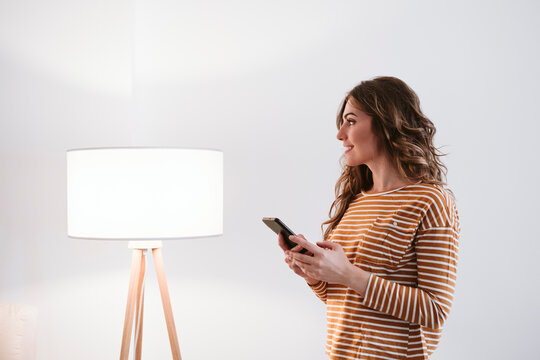Young woman holding mobile phone beside floor lamp at home