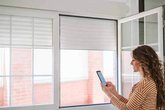 Young Woman At Home Using Mobile Phone App To Control Blinds
