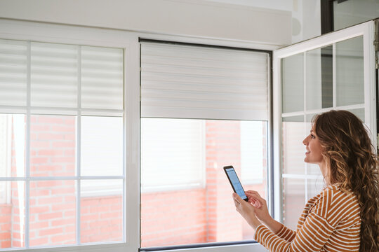 Young Woman At Home Using Mobile Phone App To Control Blinds