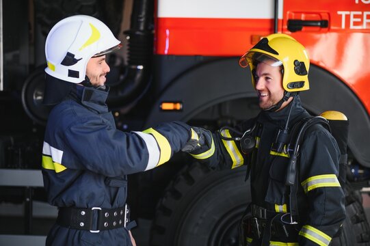 Two Firefighters In Protective Clothing In Helmets With Fire Engine, Friendly Handshake