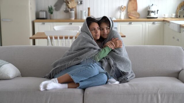 Freezing Caucasian woman and teenage girl are sits on couch wrapped in plaid due to non-working heating system in house. Young mother and daughter are trying to keep warm due to severe cold snap