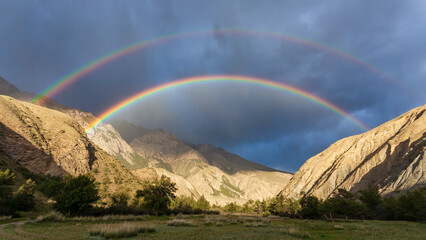 Bright full rainbow on the background of storm cloud over mountain range and valley.