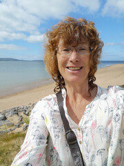 A mature woman takes a selfie after a summer swim in the sea. She is smiling at the camera and wearing a white blouse. Active senior enjoying retirement with outdoor activities and exercise.