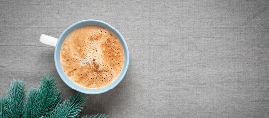 Obraz na płótnie Canvas Cup of cappuccino coffee and decorative spruce branch, gray linen texture background. Cozy Christmas background. Top view, selective focus. Banner