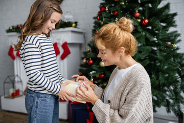 Obraz na płótnie Canvas Side view of girl giving new year present to smiling mom in knitted cardigan at home