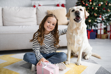 Kid holding new year present and hugging labrador on carpet in living room