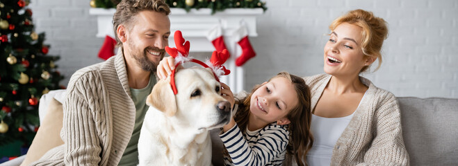 Positive family wearing christmas headband on labrador in living room, banner