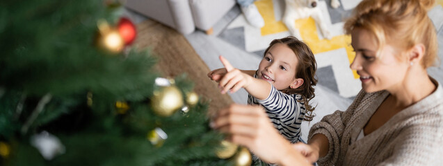 High angle view of smiling girl pointing at christmas tree near mom at home, banner