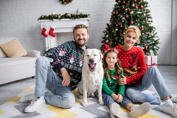 Family in christmas sweaters and jeans looking at camera near labrador at home