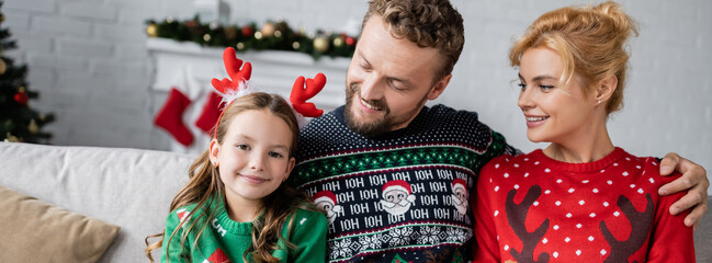 Smiling parents in sweaters looking at daughter with christmas headband, banner