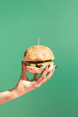 Modern scene with womans hand holding a burger against green background. Minimal composition with...