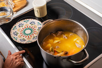 Woman's hand controlling the temperature of the ceramic hob, with a pot with stewed meat and potatoes, in the kitchen at home.