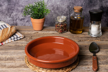 Empty rustic clay plate, next to bread, oil, pepper and salt, on a wooden table, ready to fill with food.
