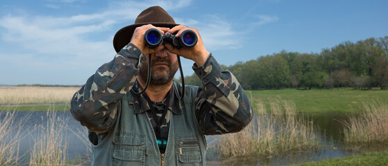 A hunter in a hat with binoculars looks out for prey against the background of the forest.
