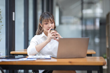  Smiling business Asian woman sitting at workplace office desk, holding a cup of coffee, she is relaxing and looking on laptop, report real estate investment data, Financial and tax systems concept.