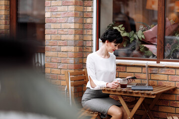 Businesswoman talking on the smartphone, working on a laptop and drinking coffee at cafe on terrace in urban city. Communication with clients remote. Coffee break, business concept. Working outdoors