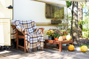 Wooden armchair near outside caravan trailer decorated for Halloween. Wooden RV house porch with garden furniture. Campsite in garden. Interior cozy yard campsite with fall flowers potted and pumpkins
