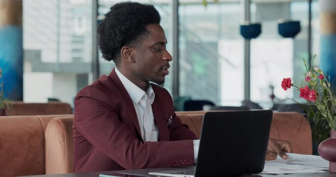 A businessman using a laptop on a wooden table in a coffee shop with a cup of coffee. He is reviewing documents created as part of a project, sorting through piques to give to boss.