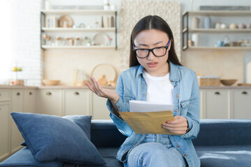 Bad letter from bank, woman sitting at home disappointed and sad reading message bad news, asian woman in kitchen.