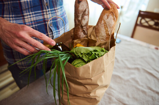 Overhead View Of A Guy Sorting Organic Fresh Vegetables While Unpacking Grocery Shopping Bag In The Kitchen At Home. Food Delivery. Healthy Grocery Purchasing In Farmers Markets And Stores