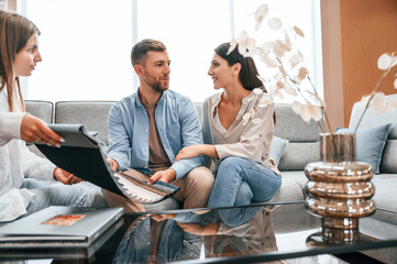 Looking at the catalogue. Woman helping couple with choosing the sofa and bed in the store