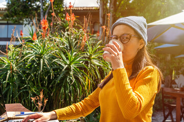 Young hipster latin woman sitting at a wooden table outside while drinking a hot beverage on a...