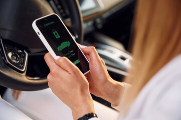 Sitting inside with smartphone. Young woman in white clothes is with her electric car at daytime