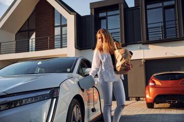 Holding shopping bag. Young woman in white clothes is with her electric car at daytime
