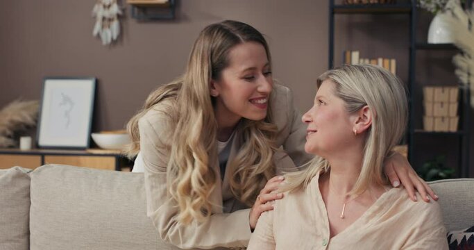 Daughter stands behind couch embraces elderly woman. The girl makes a wish, greets her mother kisses her on the cheek. Motherly love.