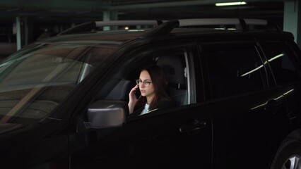 Fototapeta na wymiar A young woman in a suit speaks on the phone while sitting in a car in the parking lot. Businesswoman in glasses communicates on the phone.