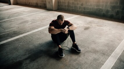 Athletic Caucasian man holding a jump rope resting on the ground in a parking lot after a workout