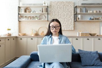 Young asian businesswoman working remotely from home sitting on sofa serious and focused using laptop for online freelance work.