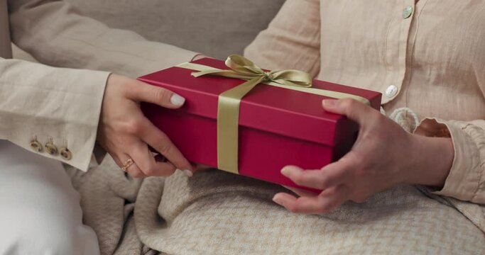 A close-up of the women's hands and the gift box. Mom opens a gift she got for daughter's birthday, senior citizen delighted with the contents of the box.