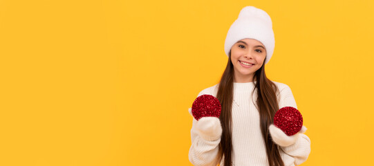 Fototapeta premium smiling child in winter hat and gloves. kid with decorative balls on yellow background. Banner of christmas child girl, studio kid winter portrait with copy space.