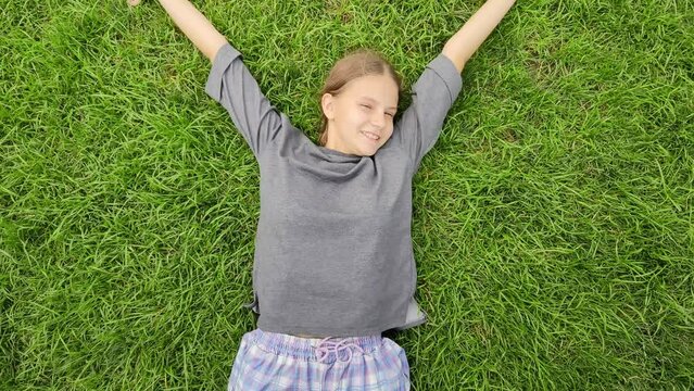 Joyful teen girl lies on grass and wave her hands imitating flapping of a bird wings. Happy child enjoy  warm summer day relax on young green grass. Wonderful mood of teenager. Adolescent psychology