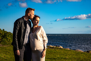 Fototapeta premium A handsome man tenderly embraces his pregnant wife and shows her love during an autumn coastal walk; autumn walk with pregnant woman; pregnant woman's belly; tenderness between partners; love