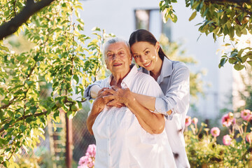 Standing in the beautiful garden and embracing each other. Young woman is with her senior mother