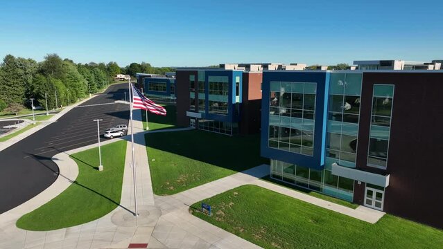 Aerial drone videography of American flag waving in front of large school building on a sunny morning. Sidewalks lead up to school building and allow students to walk. American public school theme.