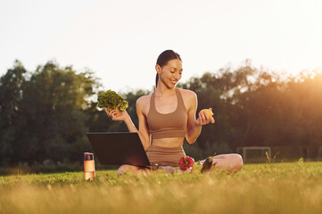 Using laptop. Holding vegetables. Young woman in yoga clothes is outdoors on the field