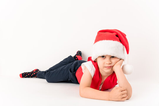 Close-up Portrait Of Lying Resentful Little Child In Red Santa Claus Hat Isolated On White Background. Beautiful Five-year European Boy. Waits A Gifts. Copy Space. Merry Christmas. No Mood. New Year