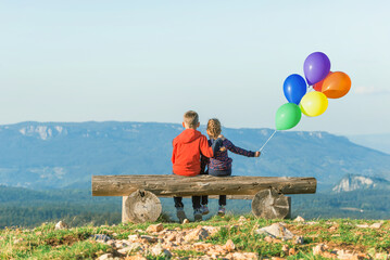 Girl and a boy are sitting on a bench with balloons swaying in the wind. View from the back....